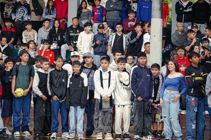 Una fila de pelados del barrio, hombro a hombro contra la reja, mirando el partido en la cancha mojada