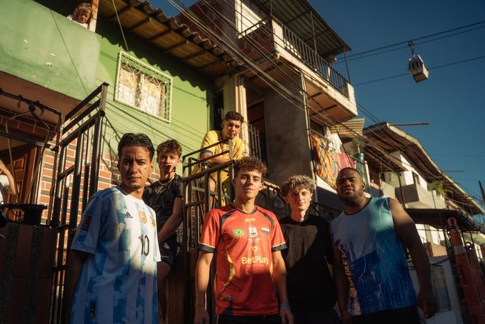 Cuatro jugadores paran en la escalera de una calle empinada del barrio, con las casas de la ladera y el metrocable al fondo