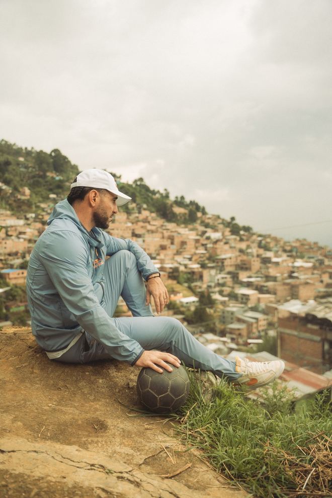 Un hombre en sudadera azul sentado en un mirador, con la mano sobre un balón y la comuna de Medellín al fondo