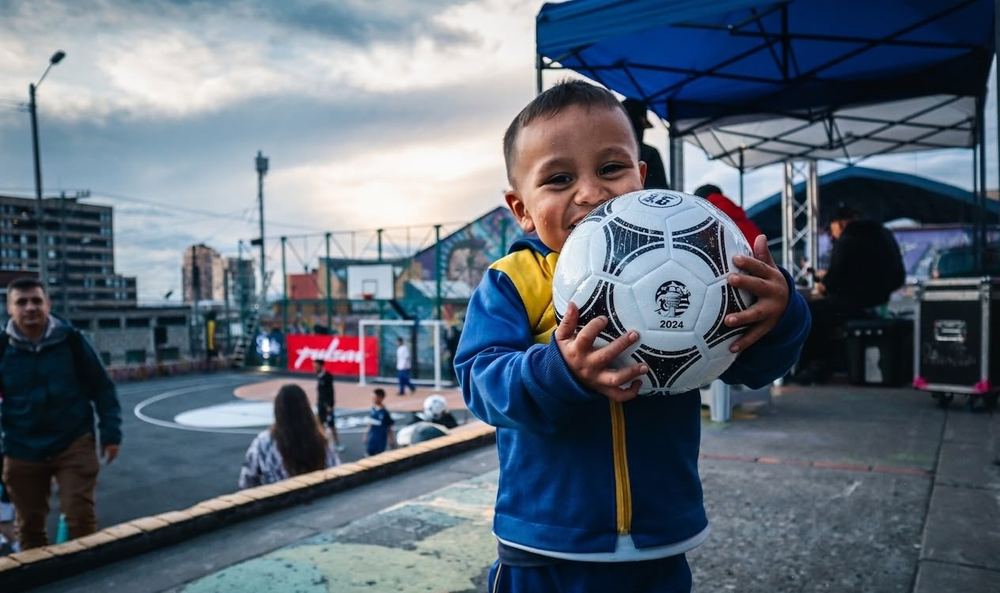 Un niño abraza el balón frente a la cancha, con Bogotá y el atardecer al fondo