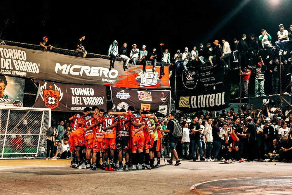 El equipo hace el círculo en la mitad de la cancha, de noche, con la gente trepada en la reja del Barrio Antioquia